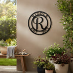 Matte black personalized letter R initial family metal sign mounted on a tan exterior porch wall above patio planters, potted greenery, and a wooden bench with a throw blanket.