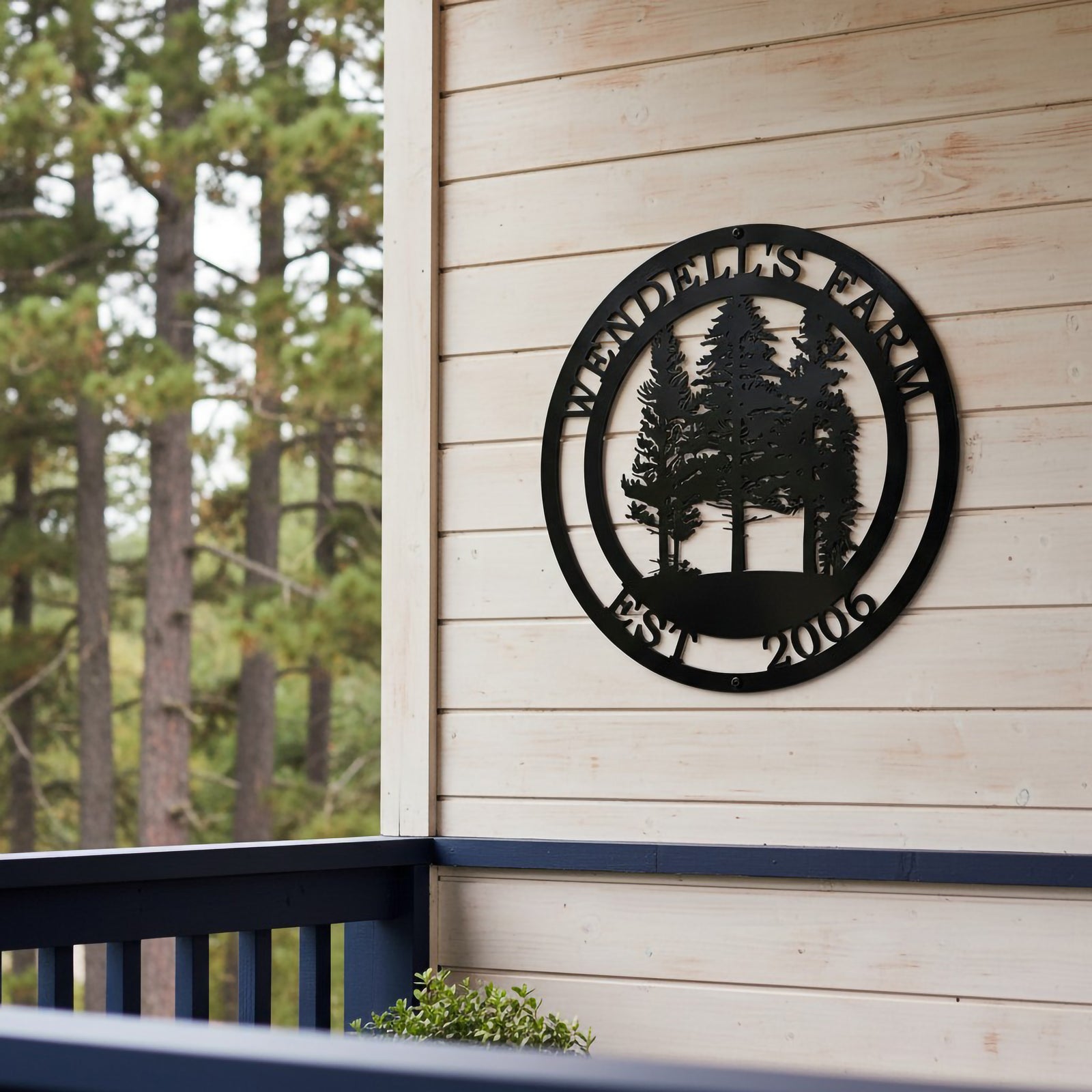 A circular black metal sign reading "Wendell's Farm Est. 2006" with a pine tree silhouette motif, mounted on a light wood siding wall above a dark blue balcony railing.