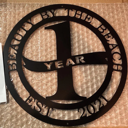 Black cutout sign with “1 YEAR” in cross shape and “BEAUTY BY THE BEACH EST 2021” encircling it, on bubble wrap.
