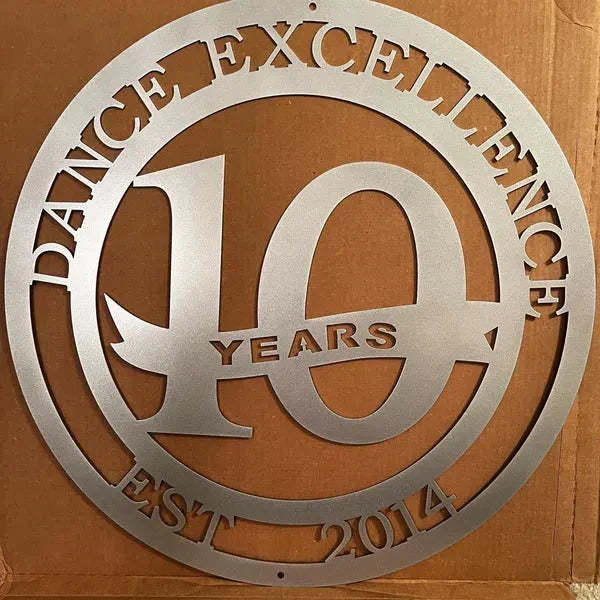 Silver circular metal sign with “DANCE EXCELLENCE,” “EST. 2014” on edge, “10 YEARS” in center, placed on cardboard.