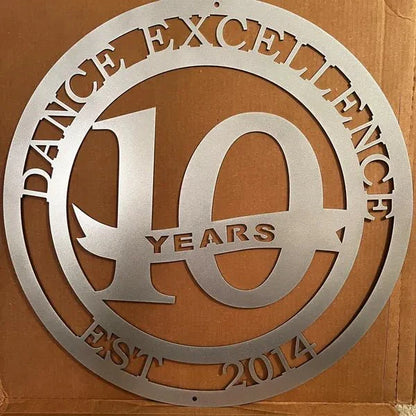 Silver circular metal sign with “DANCE EXCELLENCE,” “EST. 2014” on edge, “10 YEARS” in center, placed on cardboard.