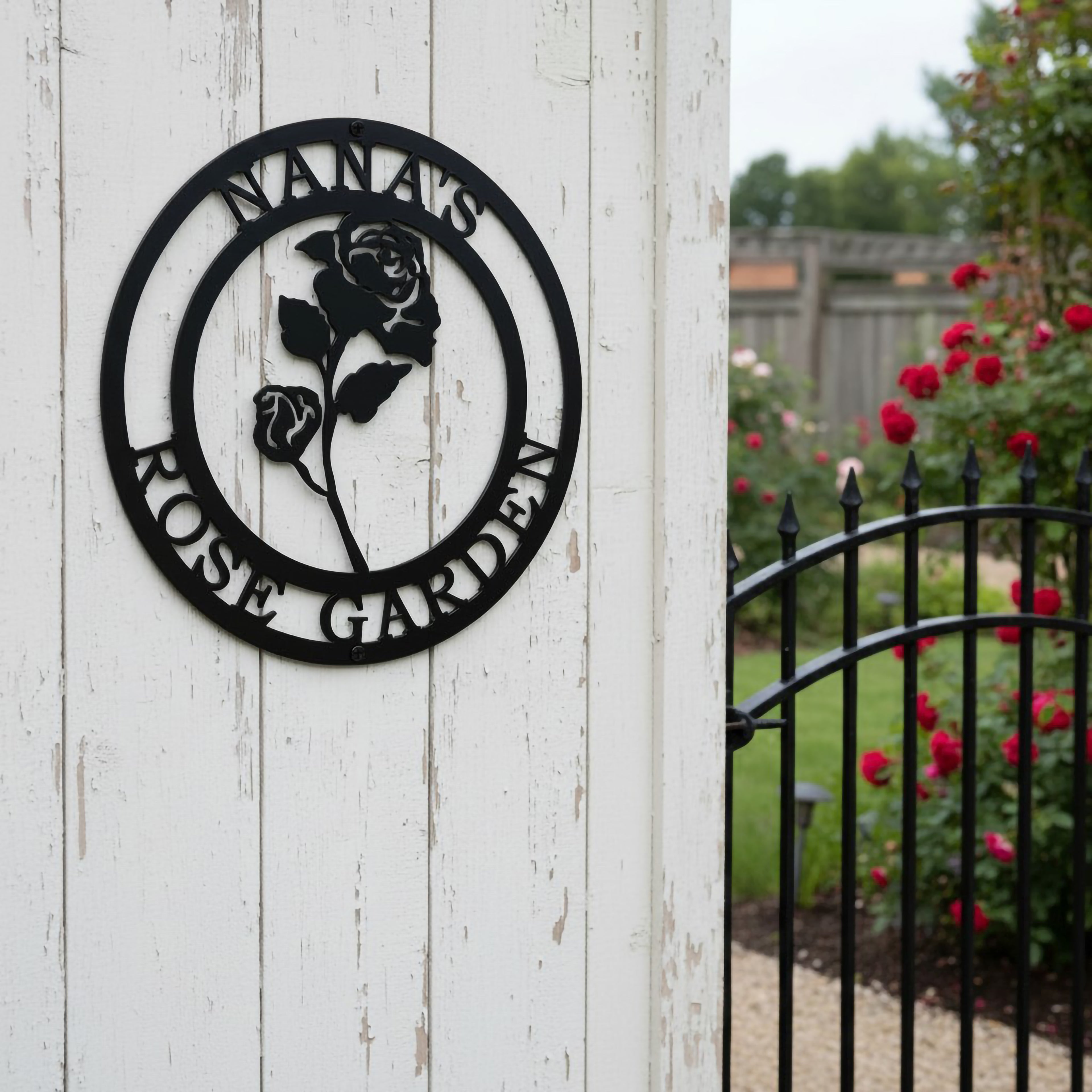 Satin black personalized botanical rose garden metal sign with custom text Nana's Rose Garden mounted on a distressed white wooden fence post next to a black wrought iron garden gate.
