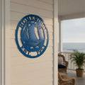 A round, laser-cut bright blue metal wall sign mounted on white vertical board-and-batten siding. The sign features a detailed sailboat riding ocean waves in the center, enclosed by a border reading "HOMEWOOD" at the top and "EST. 1953" at the bottom. A beach boardwalk porch and dark support pillar are visible in the soft-focus background.