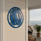 A round, laser-cut bright blue metal wall sign mounted on white vertical board-and-batten siding. The sign features a detailed sailboat riding ocean waves in the center, enclosed by a border reading "HOMEWOOD" at the top and "EST. 1953" at the bottom. A beach boardwalk porch and dark support pillar are visible in the soft-focus background.