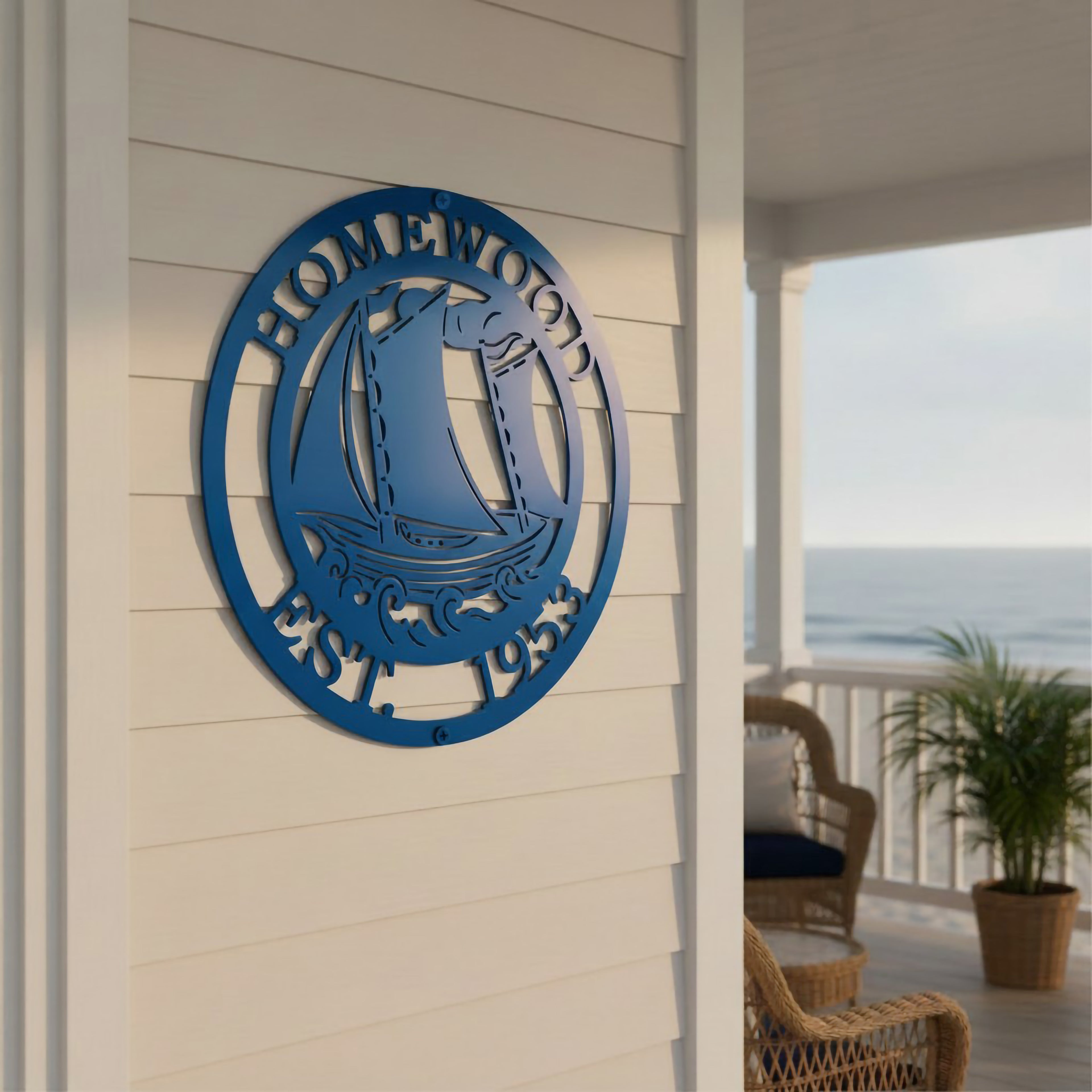 A round, laser-cut bright blue metal wall sign mounted on white vertical board-and-batten siding. The sign features a detailed sailboat riding ocean waves in the center, enclosed by a border reading "HOMEWOOD" at the top and "EST. 1953" at the bottom. A beach boardwalk porch and dark support pillar are visible in the soft-focus background.