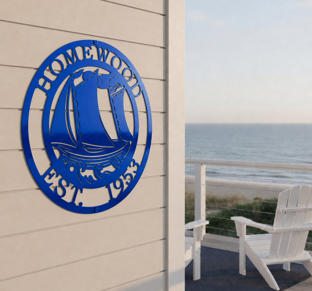 A glossy, candy cobalt blue circular metal family sign featuring a sailboat design with the text "HOMEWOOD EST. 1953". The sign is mounted onto light beige horizontal lap siding on an open deck overlooking the ocean, with a white Adirondack chair in the background.