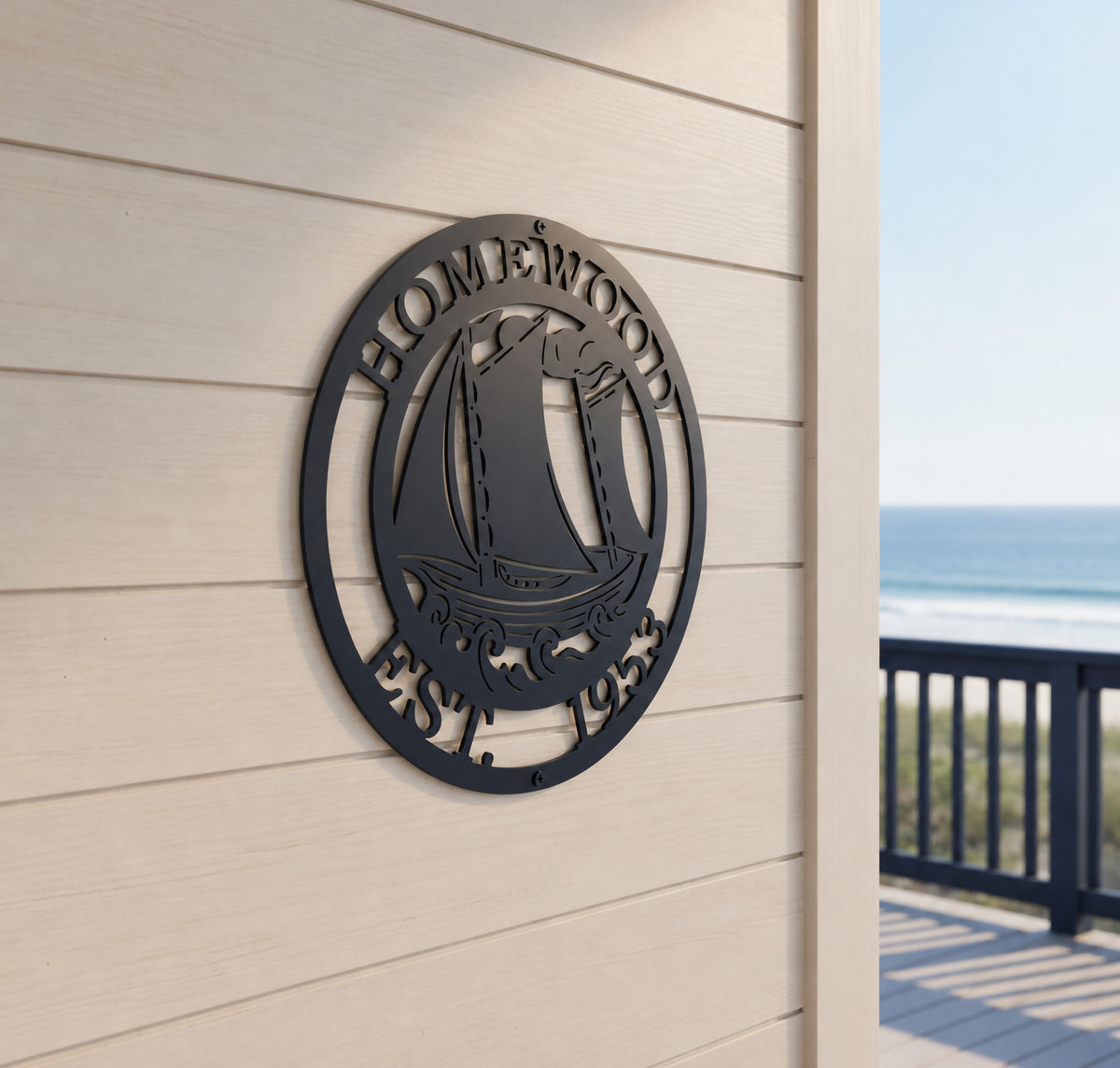 A round, dark navy blue laser-cut metal sign mounted at an angle on light beige horizontal lap siding. The sign features a stylized cutout of a sailboat on rolling ocean waves, bordered by the text "HOMEWOOD" on top and "EST. 1953" at the bottom. In the background, a dark blue wooden deck railing looks out over a sunny beach and calm ocean.