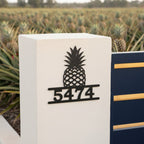 Satin black pineapple house number sign with address 5474 mounted on a white stucco gate post near a pineapple field