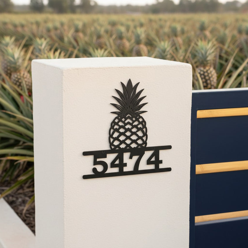 Satin black pineapple house number sign with address 5474 mounted on a white stucco gate post near a pineapple field