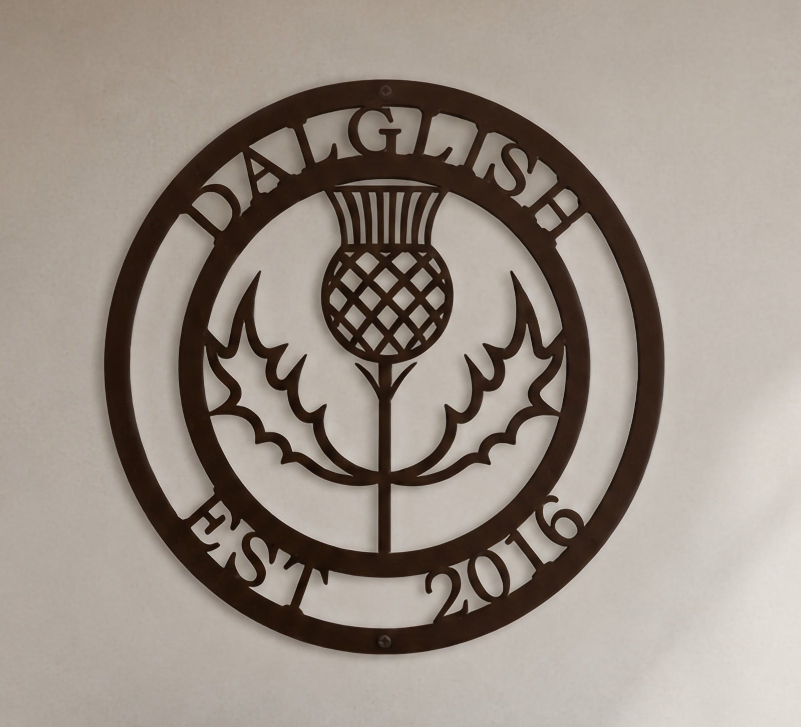 Close-up product shot of a dark espresso brown circular metal wall sign featuring a Scottish thistle flower design with custom name Dalglish and Est 2016.