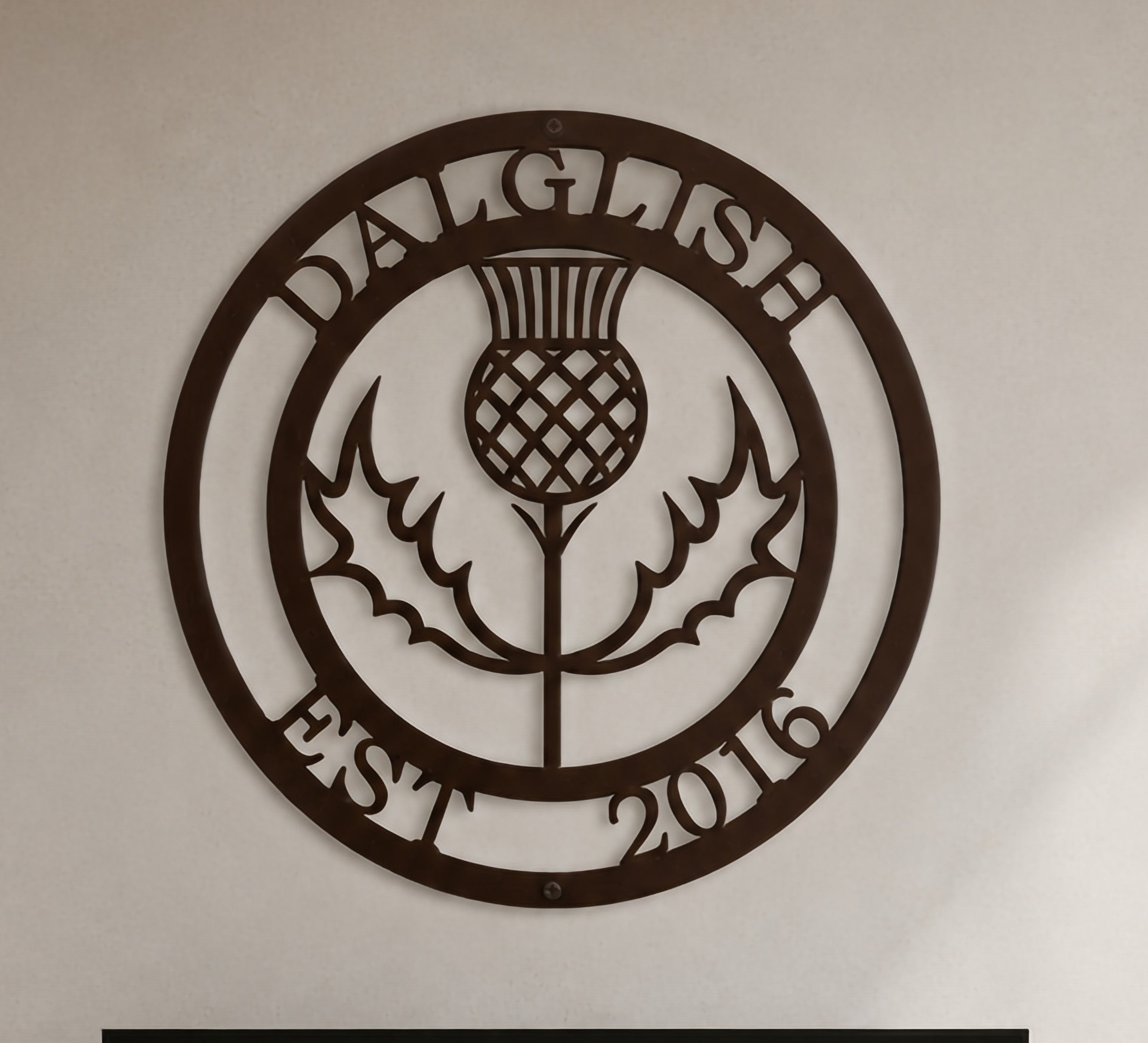 Close-up product shot of a dark espresso brown circular metal wall sign featuring a Scottish thistle flower design with custom name Dalglish and Est 2016.