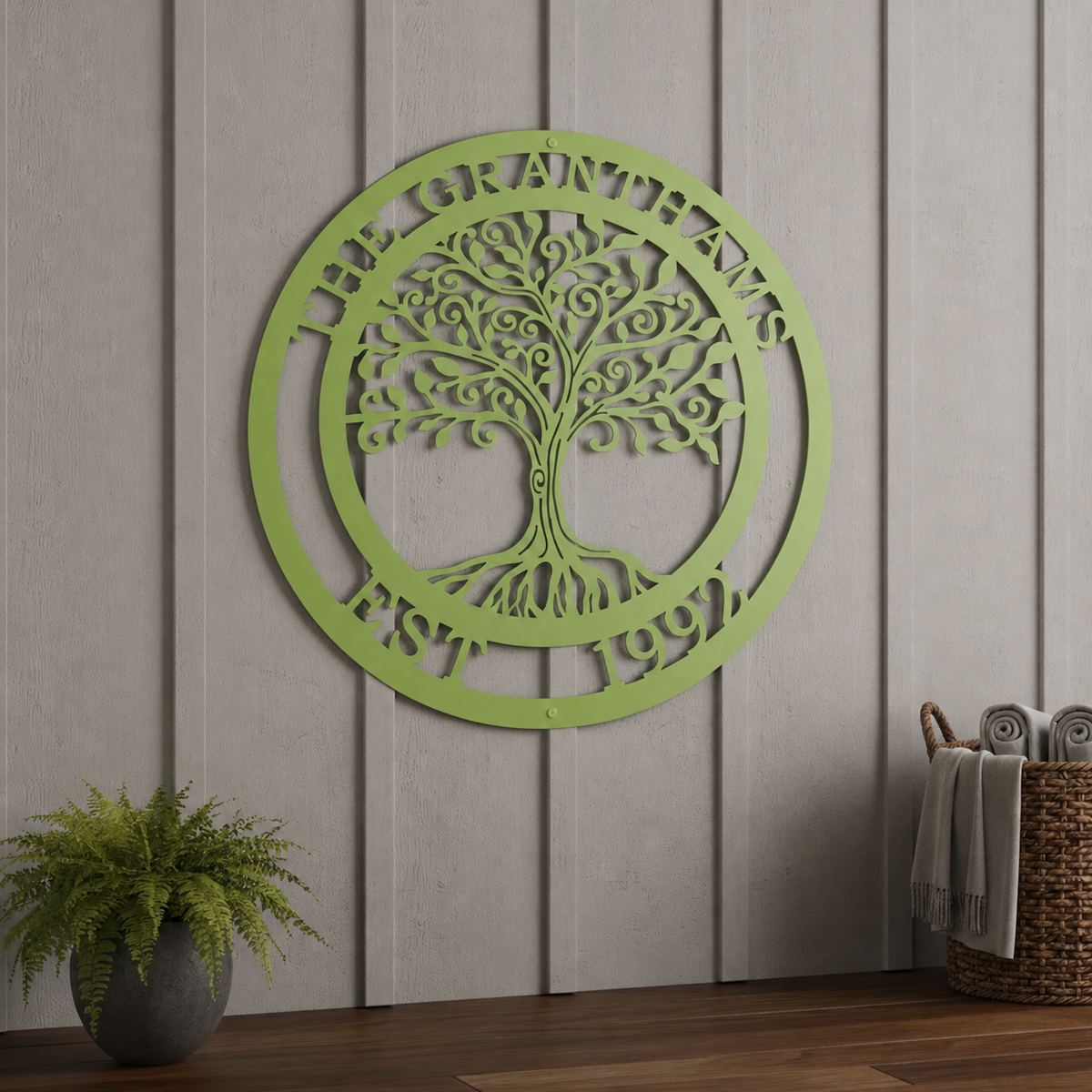 A refreshing lime green powder-coated circular metal sign featuring the text "THE GRANTHAMS EST. 1992" around a decorative tree silhouette. It is mounted indoors on a vertical slate-gray shiplap accent wall above a wooden floor, next to a potted green fern and a woven towel basket.
