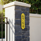 Yellow vertical house number sign with black cutout numbers displayed on a stone and stucco gate pillar