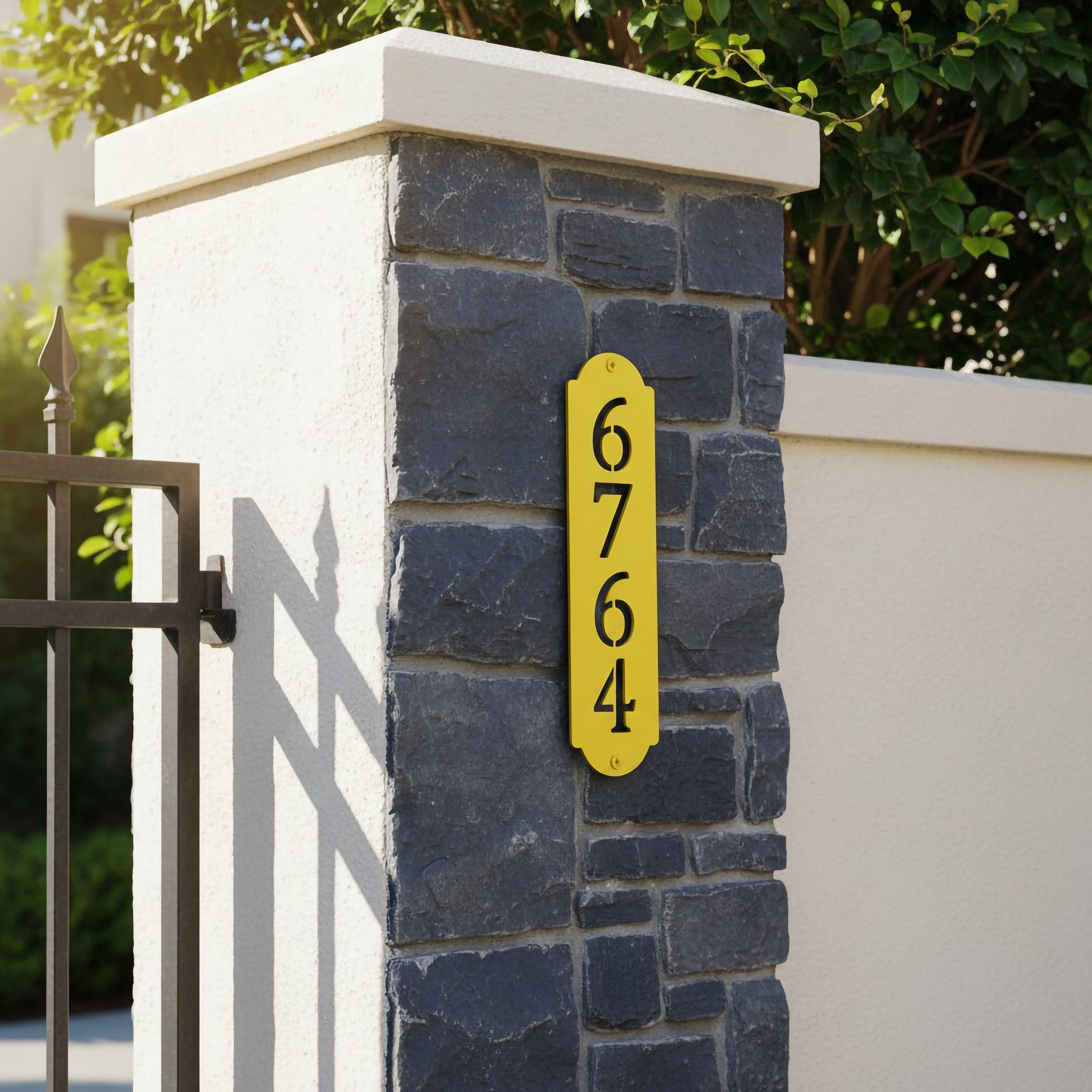 Yellow vertical house number sign with black cutout numbers displayed on a stone and stucco gate pillar