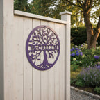 Round purple powder-coated metal tree of life sign cut with the family name McCallum mounted on a white painted wooden post fence in a garden setting.