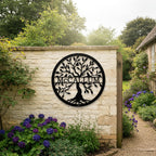 Large black round metal tree of life monogram sign featuring the name McCallum hung on a rustic cream stone wall next to an open wooden gate and a gravel garden pathway.