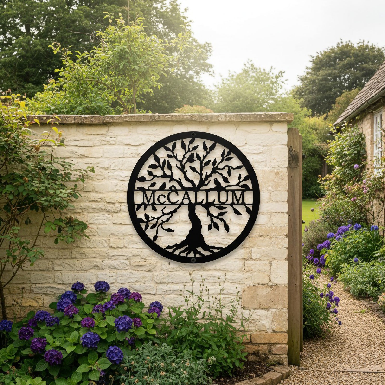 Large black round metal tree of life monogram sign featuring the name McCallum hung on a rustic cream stone wall next to an open wooden gate and a gravel garden pathway.