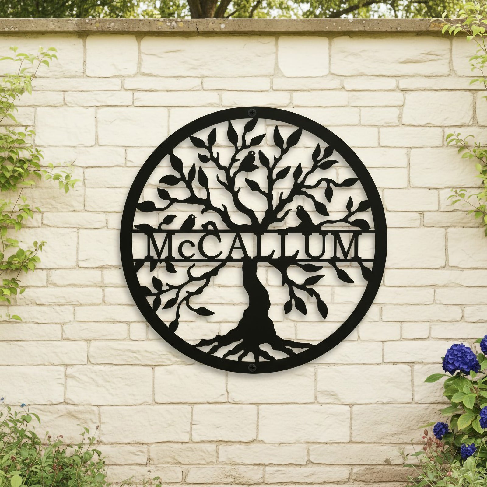 Matte black round metal tree of life sign personalized with the family surname McCallum mounted centrally on a light-colored brick stone garden wall.