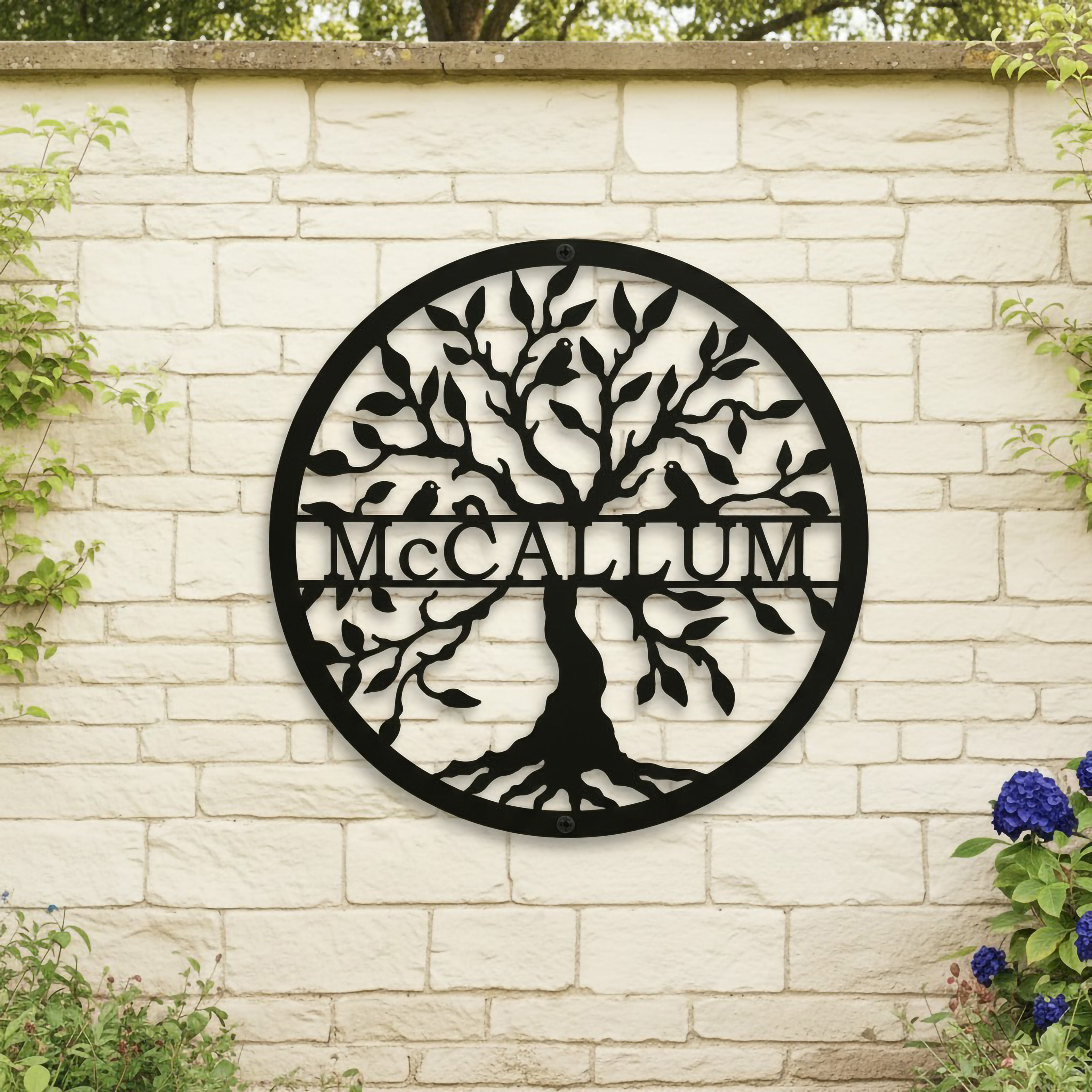 Matte black round metal tree of life sign personalized with the family surname McCallum mounted centrally on a light-colored brick stone garden wall.