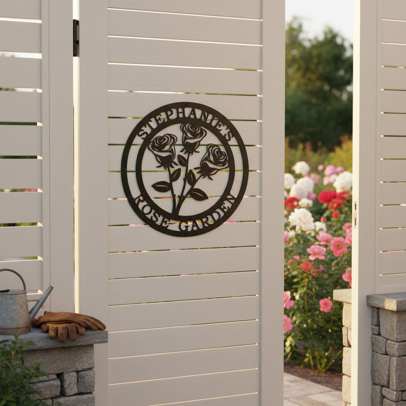 Satin black personalized botanical rose bouquet metal sign with custom text Stephanie's Rose Garden mounted centrally on a white slatted privacy gate overlooking a flower bed.