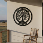 A satin black circular metal sign displaying a Tree of Life motif and the text "WAYMAKER RANCH" is mounted on a plain white stucco patio wall. A wooden rocking chair draped with a cozy blanket sits on the balcony, looking out over a wide rolling pasture where horses graze under a clear sky.