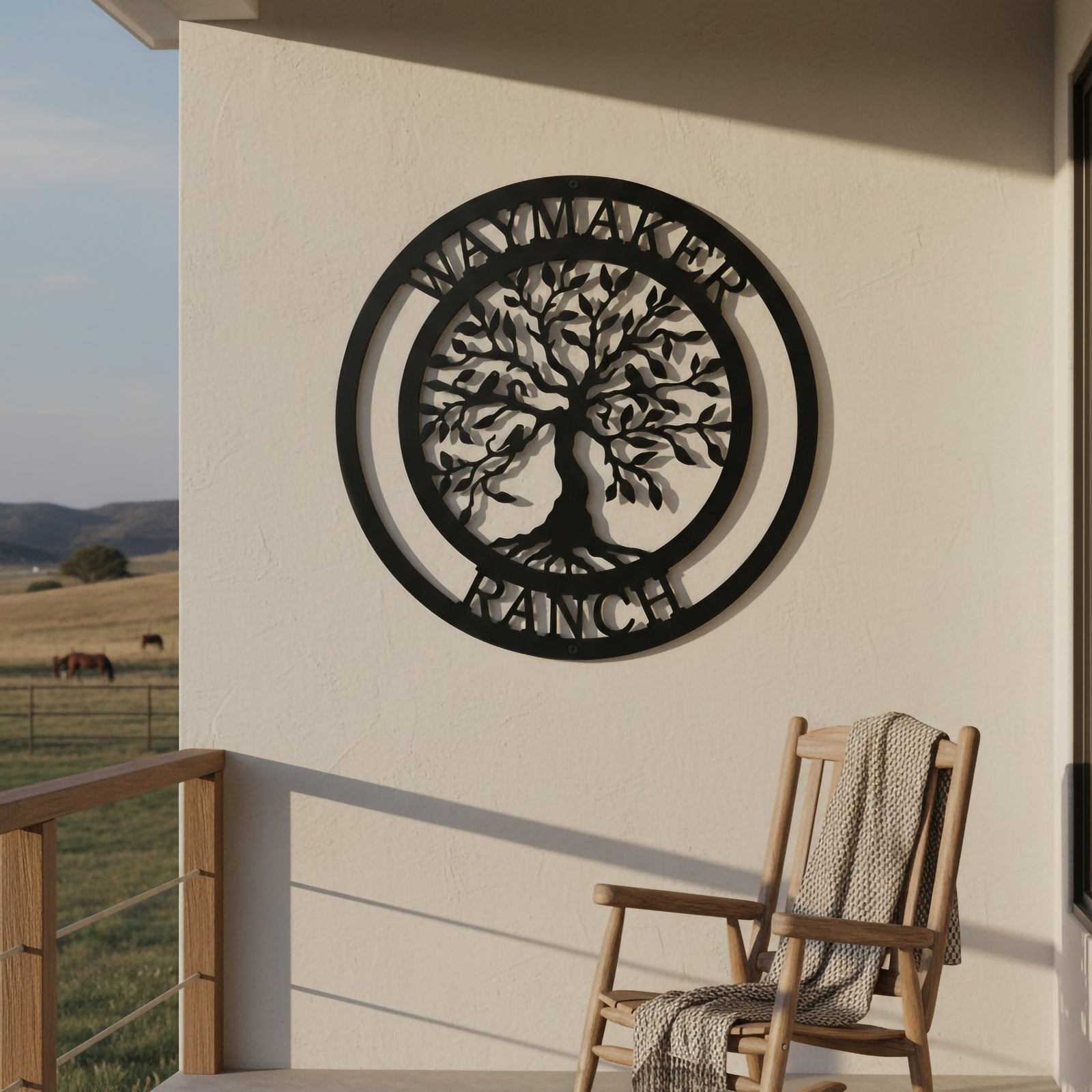 A satin black circular metal sign displaying a Tree of Life motif and the text "WAYMAKER RANCH" is mounted on a plain white stucco patio wall. A wooden rocking chair draped with a cozy blanket sits on the balcony, looking out over a wide rolling pasture where horses graze under a clear sky.
