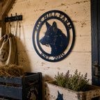 A product mockup of a black circular metal sign displaying a fox silhouette with placeholder text reading "YOUR TEXT HERE" on the top and bottom arches. The sign is displayed against a white brick wall grid with small blue flowers in white pots on a wooden ledge below.