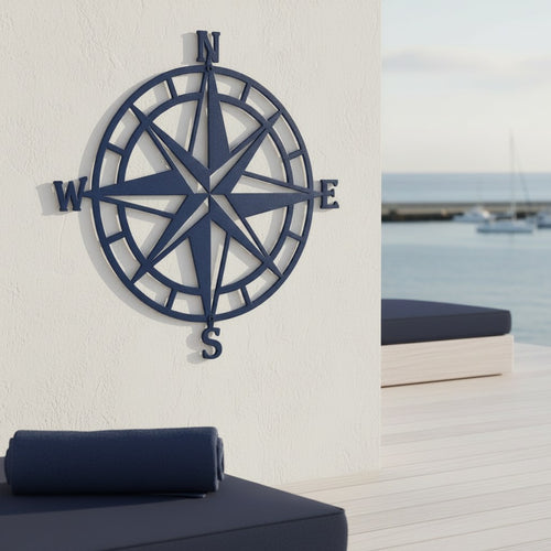 A dark navy blue metal compass rose plaque mounted on a stucco wall at a beachside resort deck, with a rolled navy towel and an ocean view in the background.