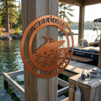A copper-colored circular metal boat sign personalized with "The Simoneaus," mounted on a natural wooden dock post overlooking a sunny lake.