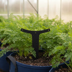 A black T-shaped metal garden stake placed in a rich soil bed surrounded by vibrant green basil plants, kale, and a potted cherry tomato plant laden with ripening fruit.