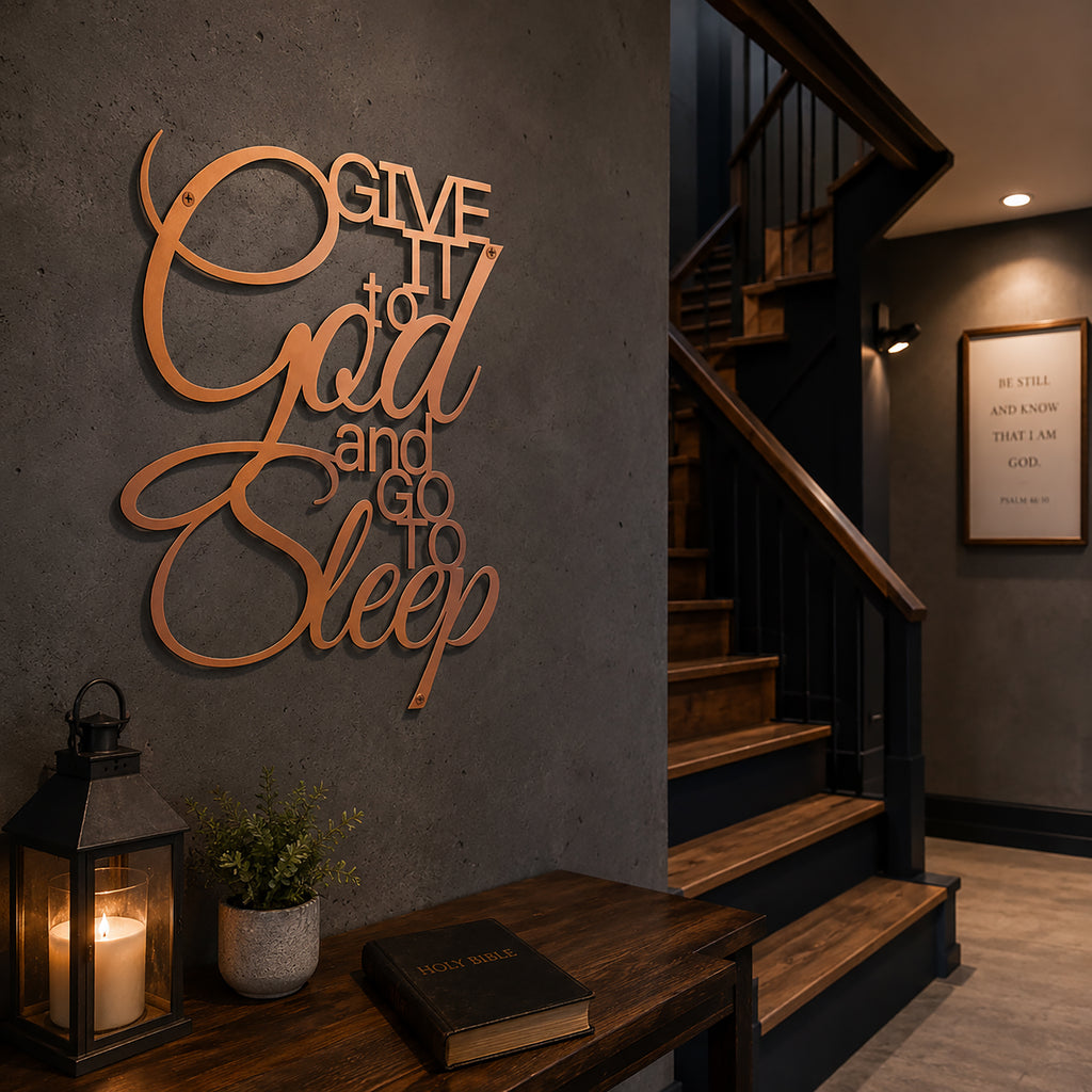 A metallic rose gold script metal sign stating "Give It to God and Go to Sleep" is mounted on a smooth dark gray wall next to a wooden staircase. A small wooden table underneath holds a Holy Bible and a black metal candle lantern illuminating the corner.