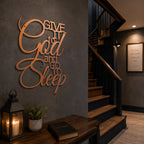 A metallic rose gold script metal sign stating "Give It to God and Go to Sleep" is mounted on a smooth dark gray wall next to a wooden staircase. A small wooden table underneath holds a Holy Bible and a black metal candle lantern illuminating the corner.