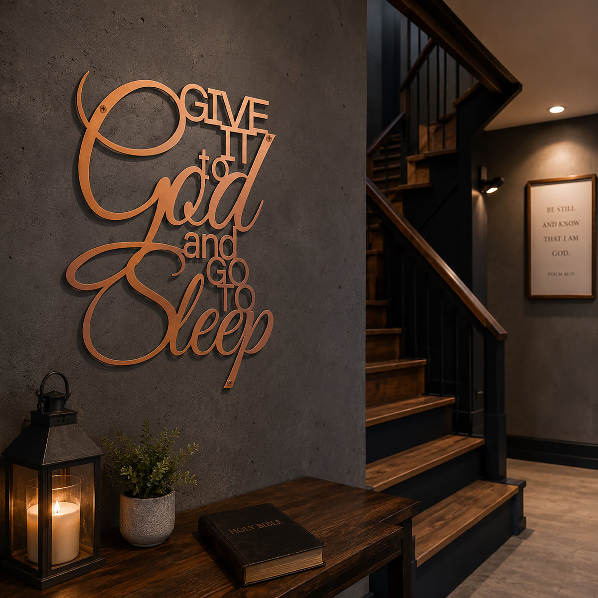 A metallic rose gold script metal sign stating "Give It to God and Go to Sleep" is mounted on a smooth dark gray wall next to a wooden staircase. A small wooden table underneath holds a Holy Bible and a black metal candle lantern illuminating the corner.