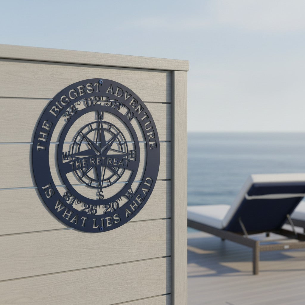 A deep navy blue circular metal compass plaque is displayed on a weathered tan wooden outdoor wall. The customized sign displays the quote "THE BIGGEST ADVENTURE IS WHAT LIES AHEAD" and "THE RETREAT." A matching navy blue cushion on a sun lounger rests on the patio deck next to the calm ocean.