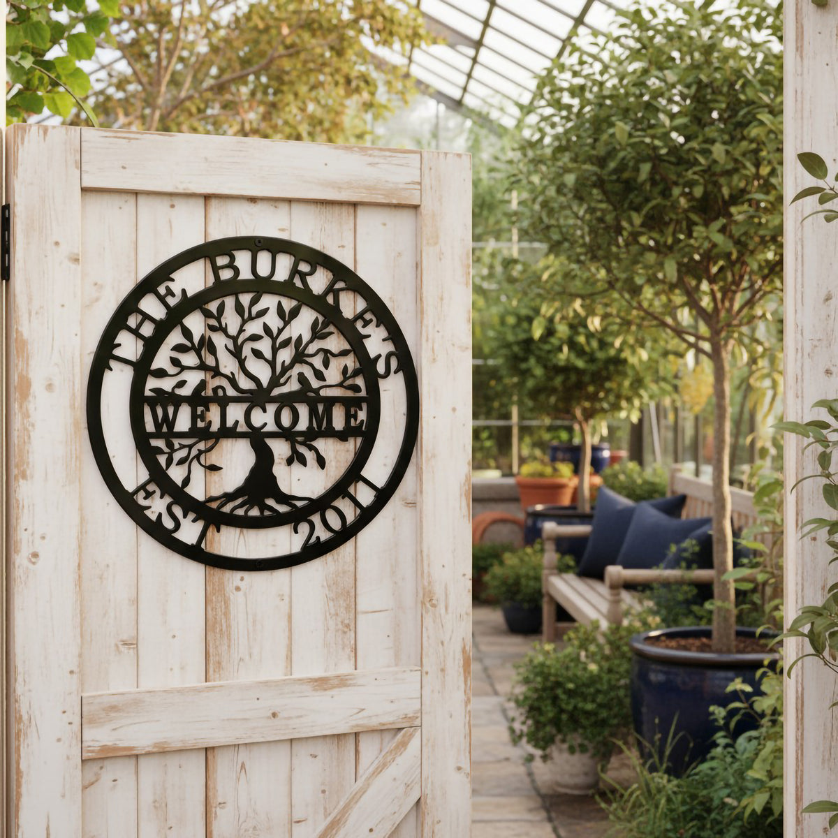  black circular metal tree sign stating "The Burkes Welcome Est. 2011," mounted on a rustic, weathered white wooden door that opens into a bright glass greenhouse filled with plants.
