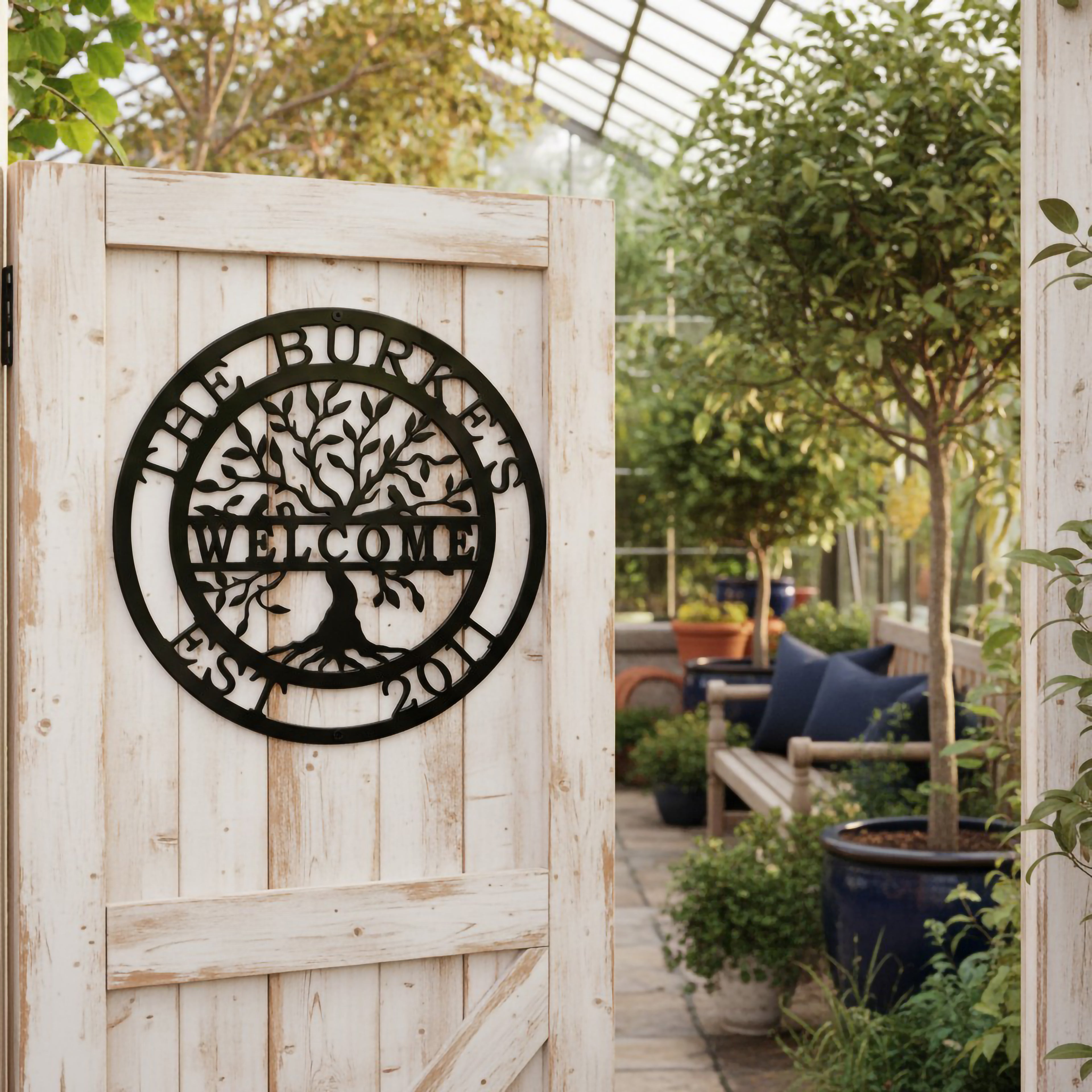  black circular metal tree sign stating "The Burkes Welcome Est. 2011," mounted on a rustic, weathered white wooden door that opens into a bright glass greenhouse filled with plants.