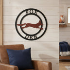 Personalized fox den metal wall sign with black round frame and rust fox design mounted on a porch wall overlooking the woods