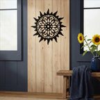 A matte black circular metal sun plaque mounted on a vertical natural wood panel, flanked by dark blue walls and a rustic wooden bench holding sunflowers in a blue vase.