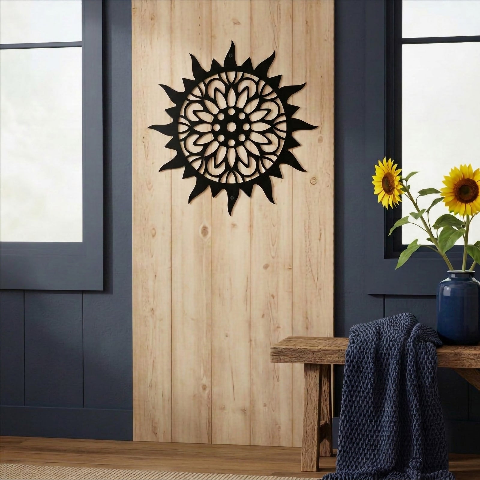 A matte black circular metal sun plaque mounted on a vertical natural wood panel, flanked by dark blue walls and a rustic wooden bench holding sunflowers in a blue vase.