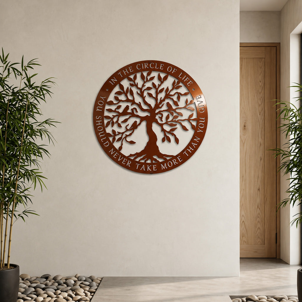 A circular metal sign with a shiny metallic Tuscan copper finish depicting a detailed Tree of Life cutout. The outer ring is engraved with an inspirational circle of life quote. It is mounted on a large off-white wall in a modern room with a light wood door, an indoor plant, and a river stone floor border.