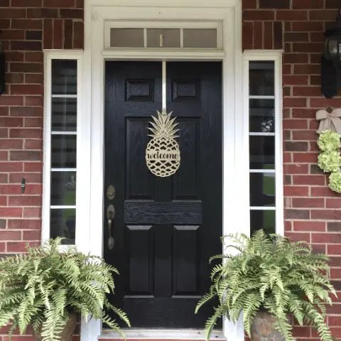 Gold pineapple welcome door decor displayed on black front door with brick porch and green plants
