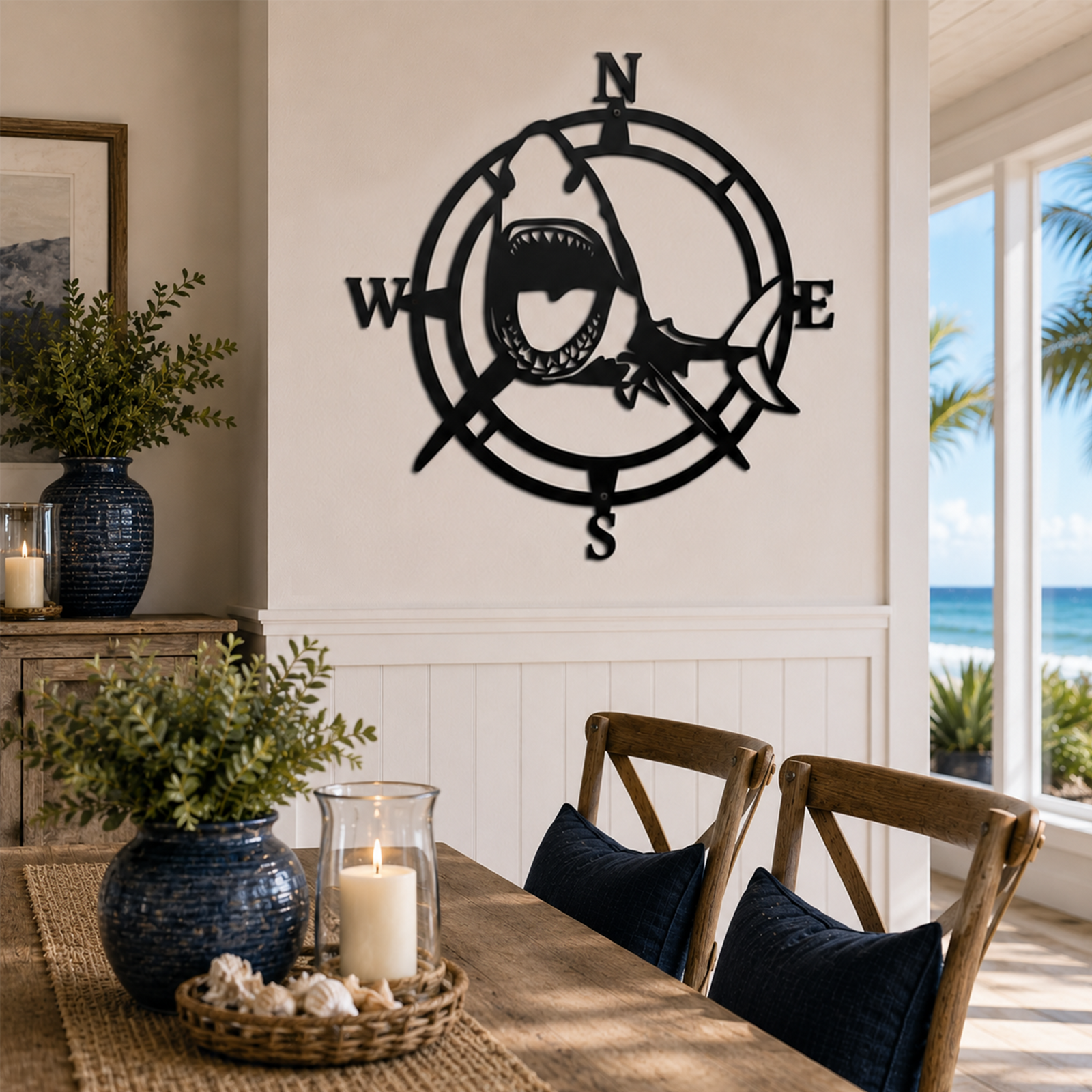 A circular black metal cutout wall art piece featuring an aggressive shark swimming through a nautical compass frame. The art is centered on a plain white wall above a rustic wooden dining table decorated with candles and ocean-themed vases, looking out a large window to a blue sea.