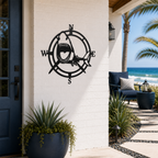 A round, laser-cut matte black metal sign depicting a striking open-mouthed great white shark positioned over a traditional nautical compass with N, S, E, and W directional markers. The sign is mounted on a white brick entryway wall next to a dark blue front door, with a sunny ocean beach walkway visible in the background.