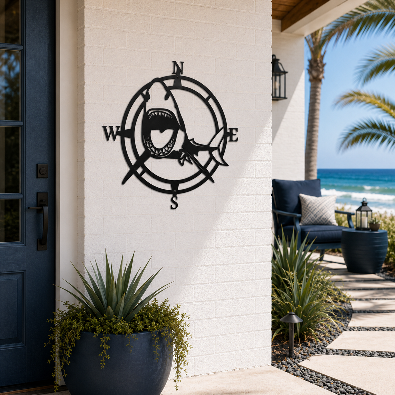 A round, laser-cut matte black metal sign depicting a striking open-mouthed great white shark positioned over a traditional nautical compass with N, S, E, and W directional markers. The sign is mounted on a white brick entryway wall next to a dark blue front door, with a sunny ocean beach walkway visible in the background.