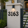 Solid black traditional-style house numbers showing "3163" mounted on a concrete pillar at a property entrance. An overhead light accentuates the numbers, which are framed by a dark vertical bar fence on the left and a matching secure entrance gate on the right.