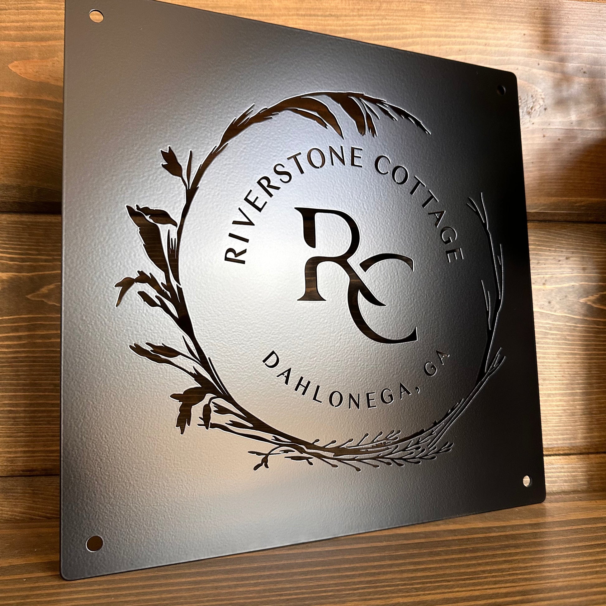 Laser-cut steel sign with “RIVERSTONE COTTAGE,” “DAHLONEGA, GA,” and “RC” in center, with decorative leaf design.