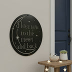 Satin black circular metal cutout sign with "I love you to the moon and back" text mounted on a plain wall next to a wooden stool decorated with star lights.