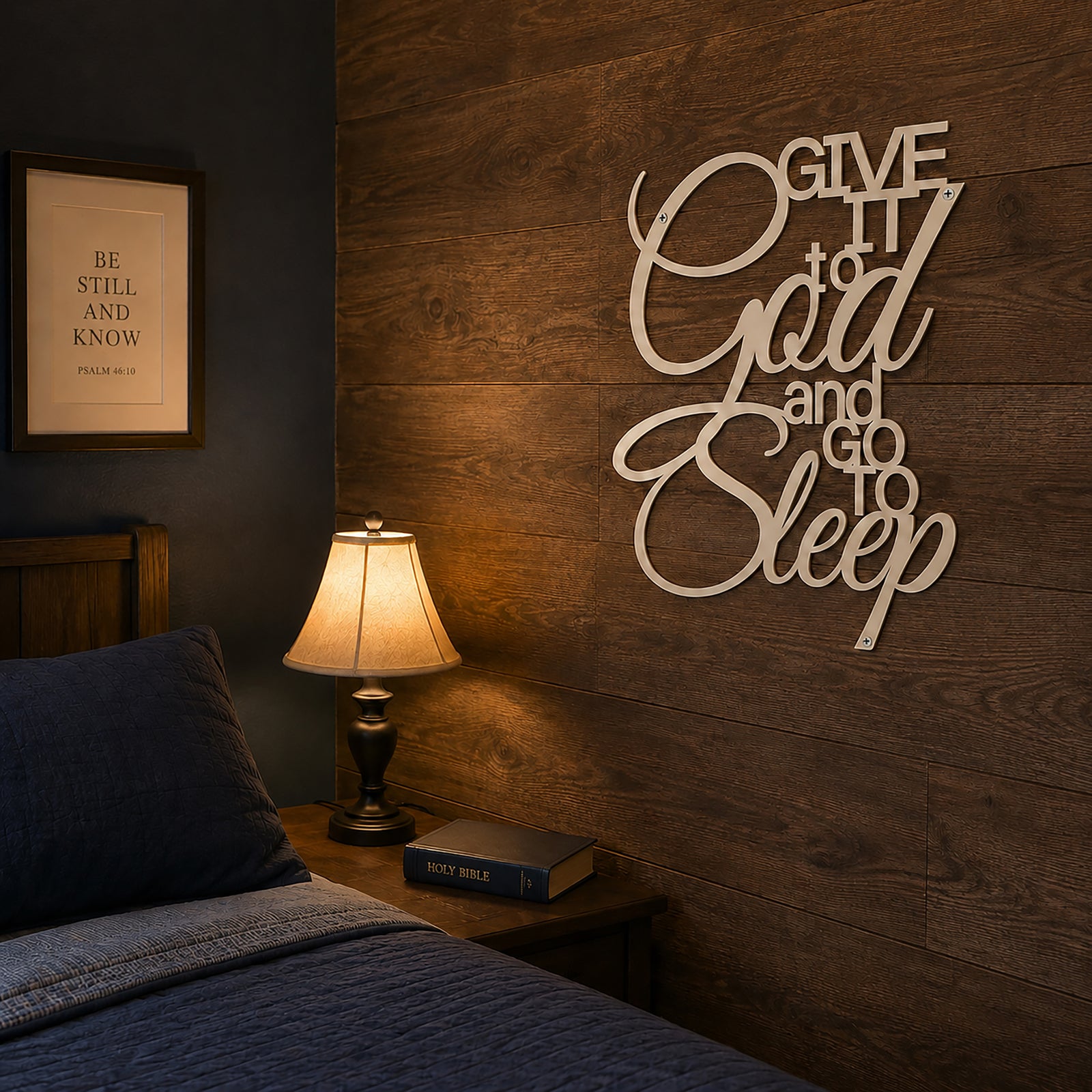A cream ivory colored metal typographic sign reading "Give It to God and Go to Sleep" is mounted on a rustic, dark brown wood-paneled bedroom wall. The sign hangs above a wooden nightstand displaying a closed Holy Bible and a small lamp casting a warm glow near a bed with a dark blue comforter.