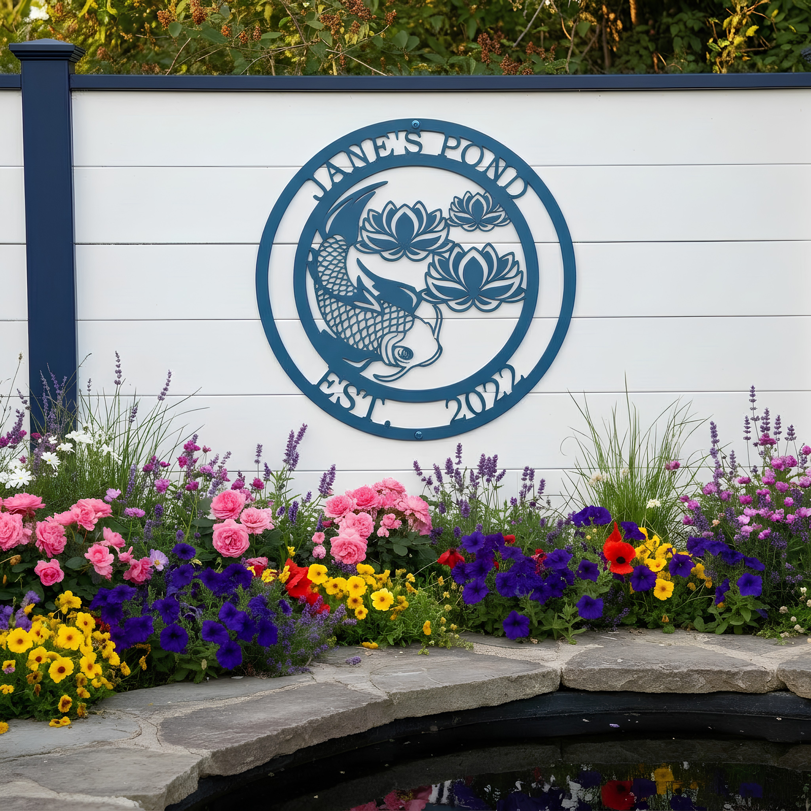 A large teal-blue metal round sign stating "Jane's Pond Est. 2022" with a koi fish cutout, mounted on a white privacy fence behind a lush bed of colorful flowers and stone edging.