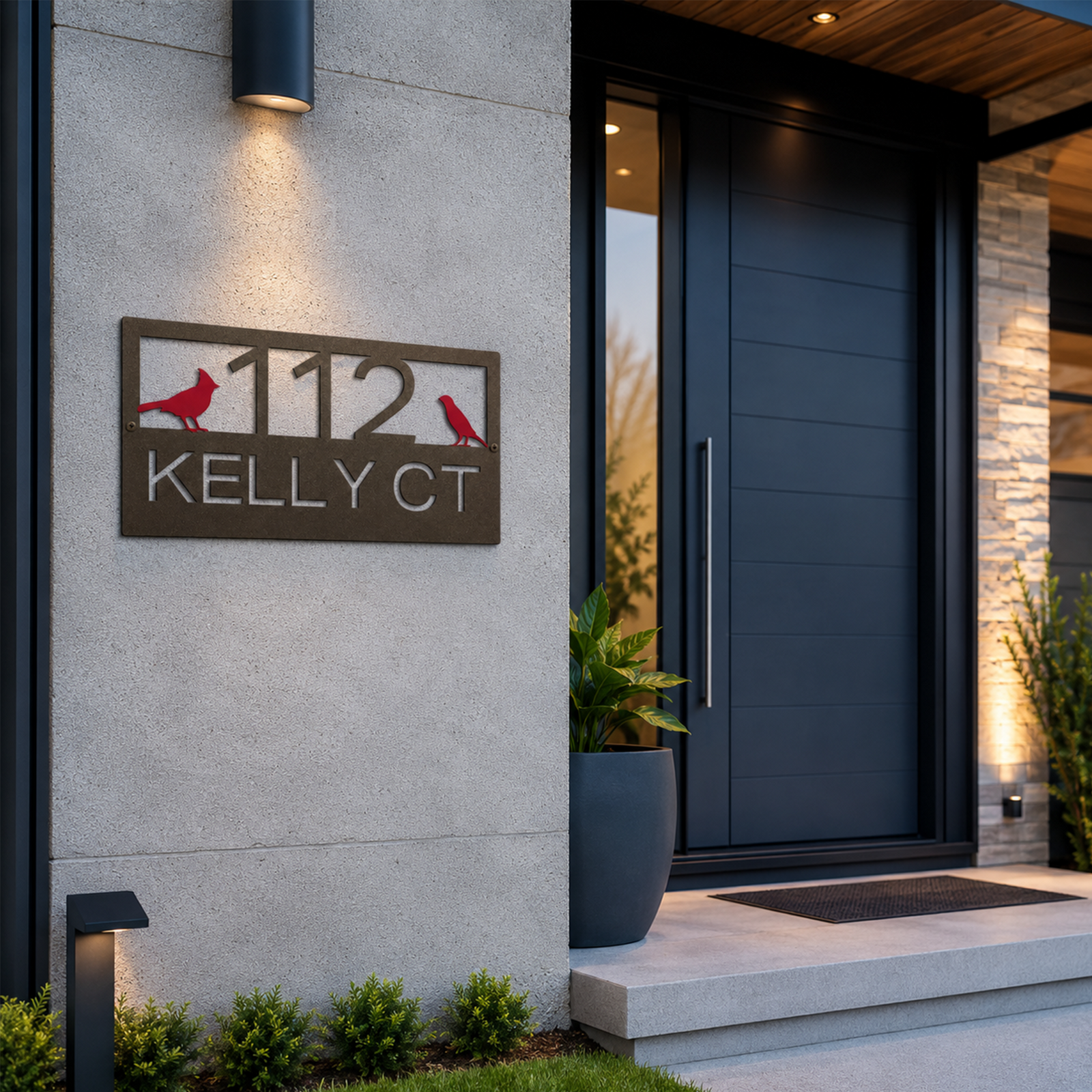 A custom rectangular address sign with a weathered bronze finish is installed on a light gray concrete wall. The sign reads "112 KELLY CT" with a pair of vibrant red birds flanking the house number. A warm overhead light illuminates the plaque next to a modern dark paneled entryway.